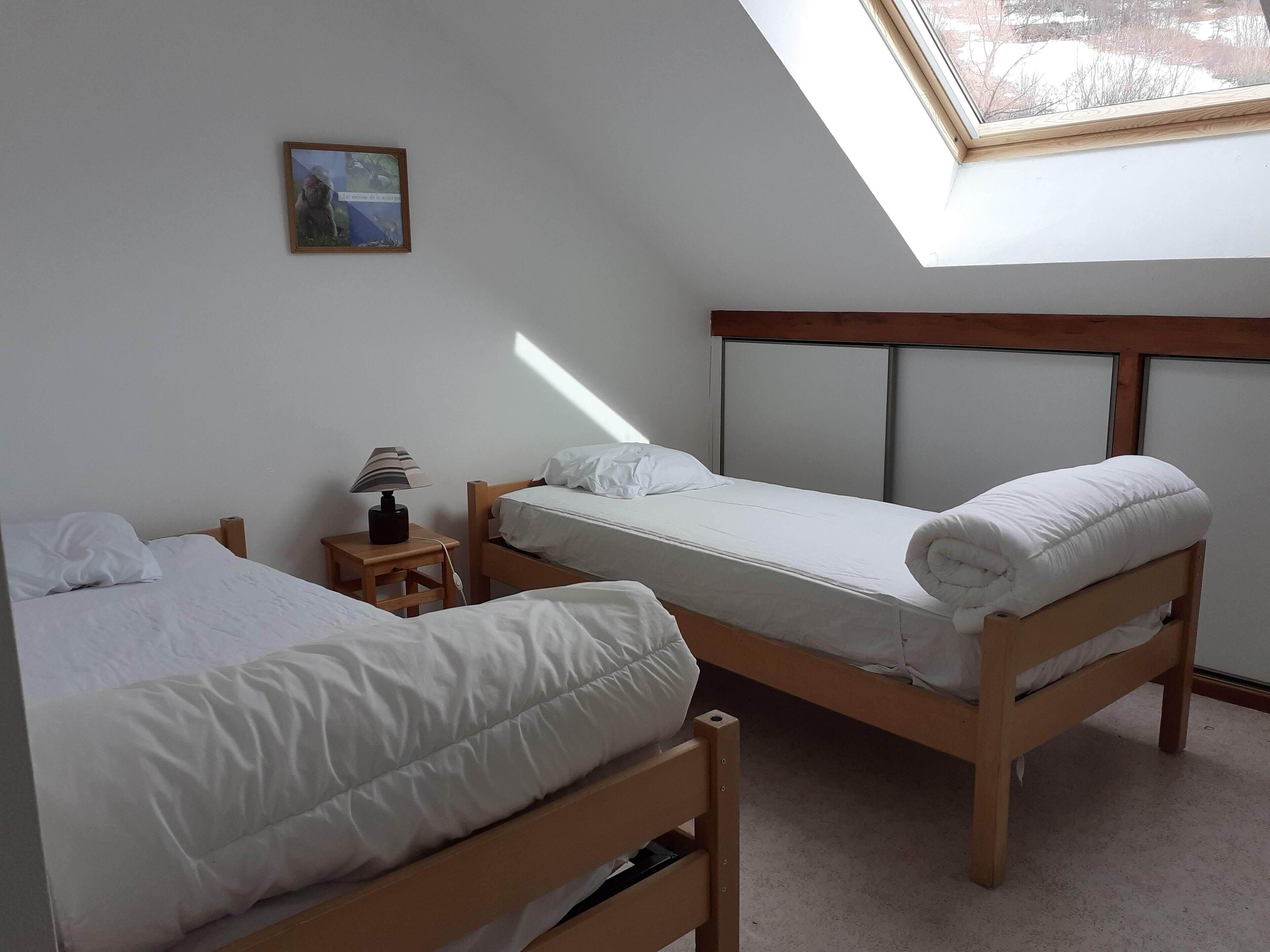 A bedroom with two wooden beds, a nightstand with a lamp, a small table, and a skylight.
