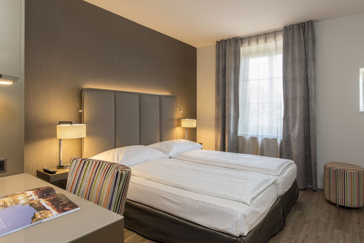 A hotel room with a double bed, two bedside lamps, a striped chair, a small table with a magazine, and a window with curtains.