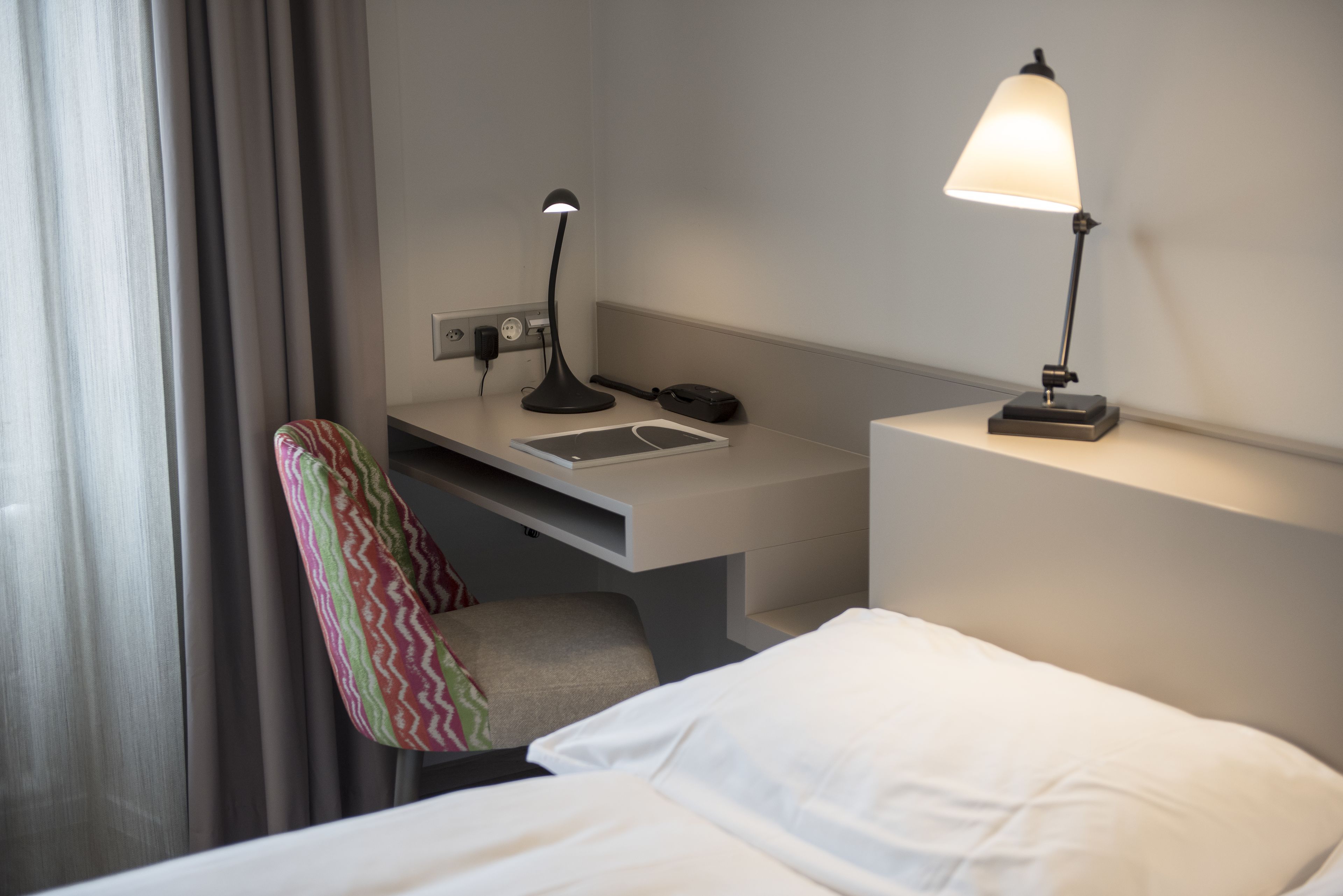 Une chambre d’hôtel avec un lit, un bureau avec une lampe, une chaise et une fenêtre avec des rideaux.