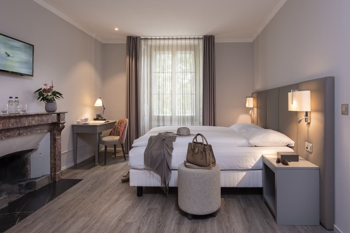 A hotel room with a bed, bedside lamps, a nightstand, a small table with a lamp, a chair, a potted plant, and a fireplace.