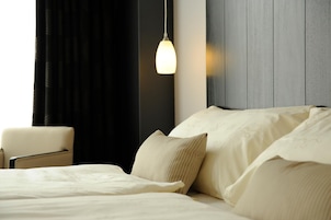 Una cama bien hecha con una colcha blanca y almohadas, una lámpara colgante, una silla y un cabecero de madera oscura.