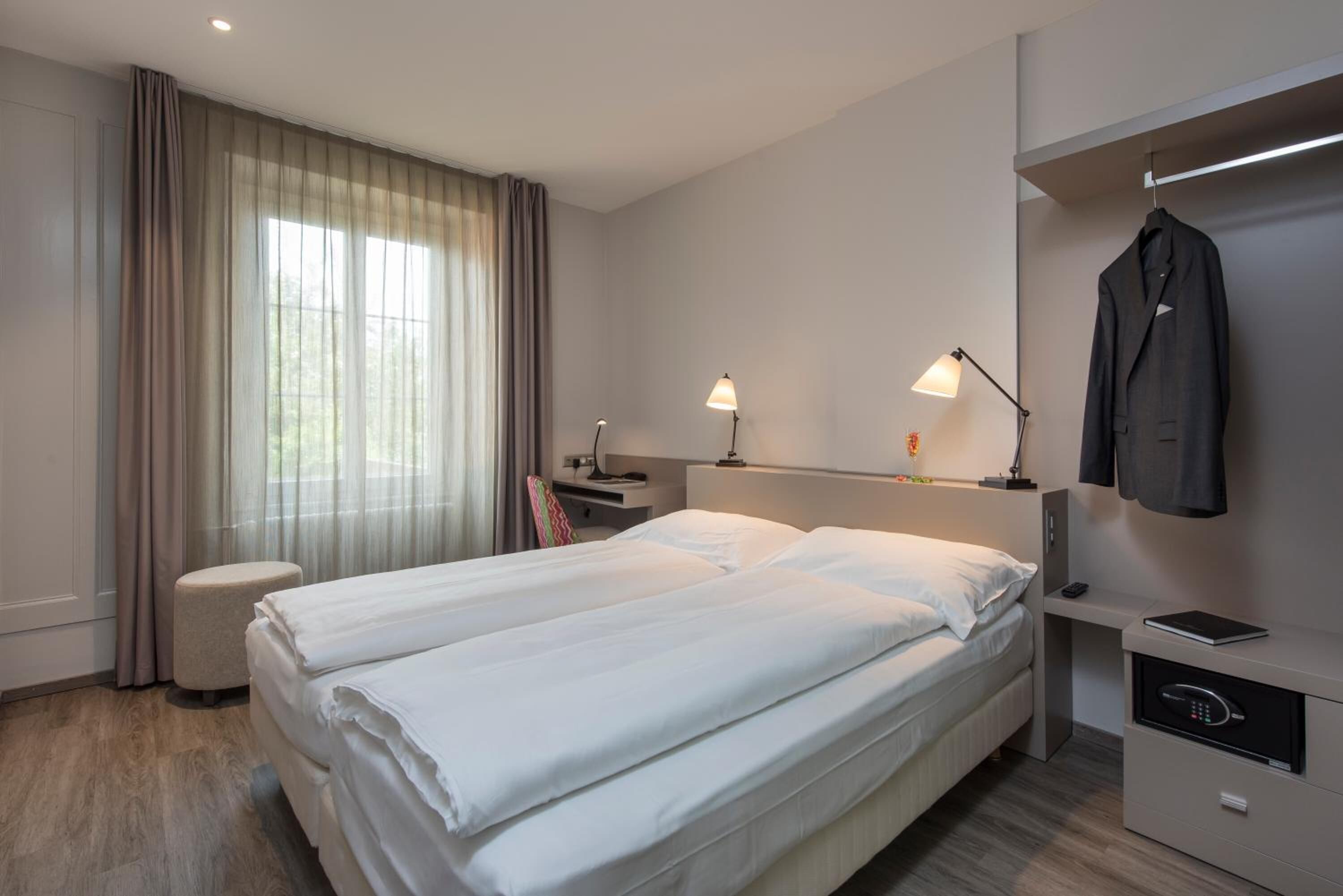 Ein Hotelzimmer mit zwei Betten, einem Schreibtisch, einem Stuhl, einer Lampe, einem Kleiderhaken und einem Safe.