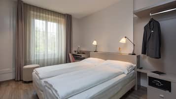 Ein Hotelzimmer mit zwei Betten, einem Schreibtisch, einem Stuhl, einer Lampe, einem Kleiderhaken und einem Safe.