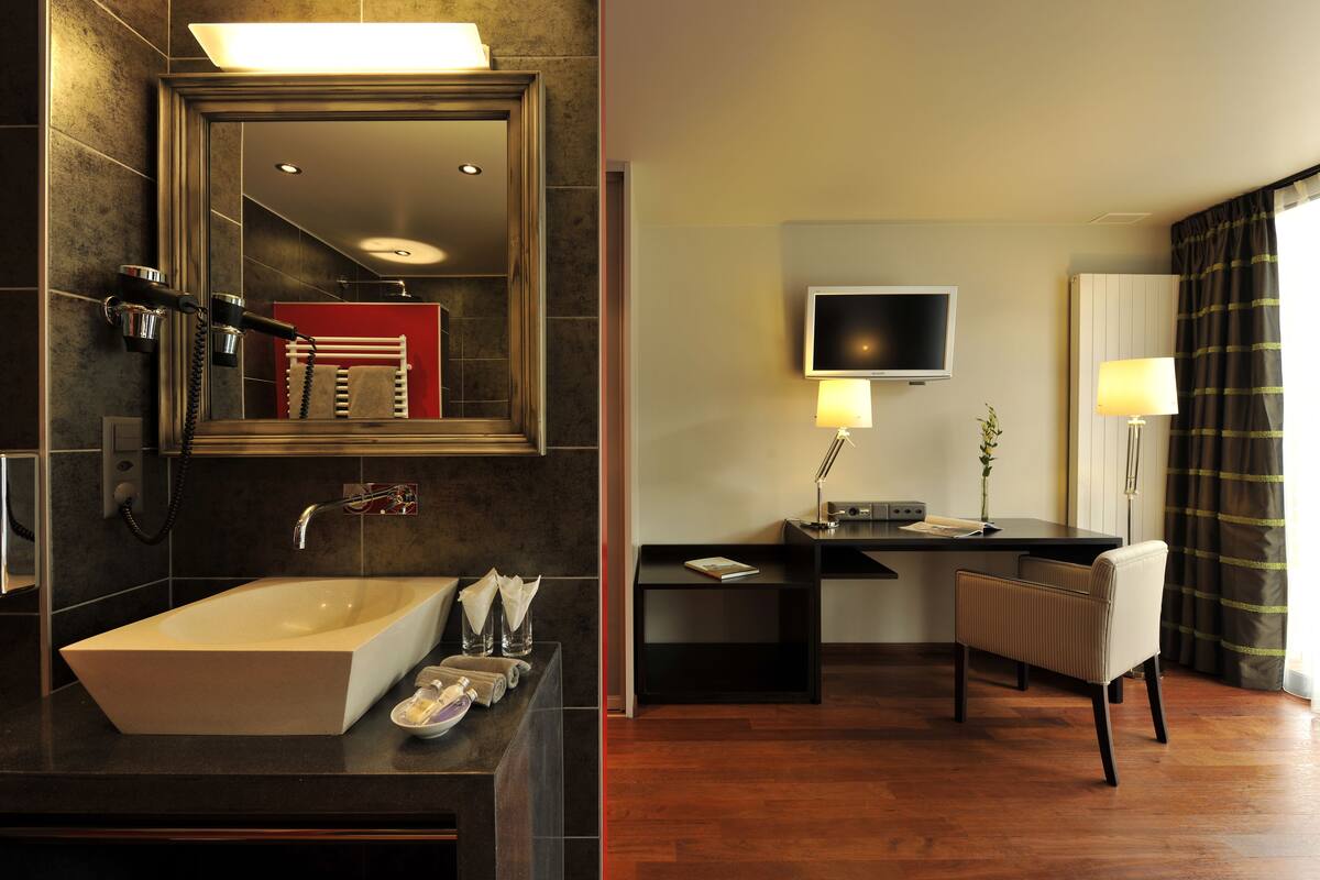 A modern bathroom with a large sink, mirror, and hairdryer. There is a desk with a chair and a TV in the adjacent room.