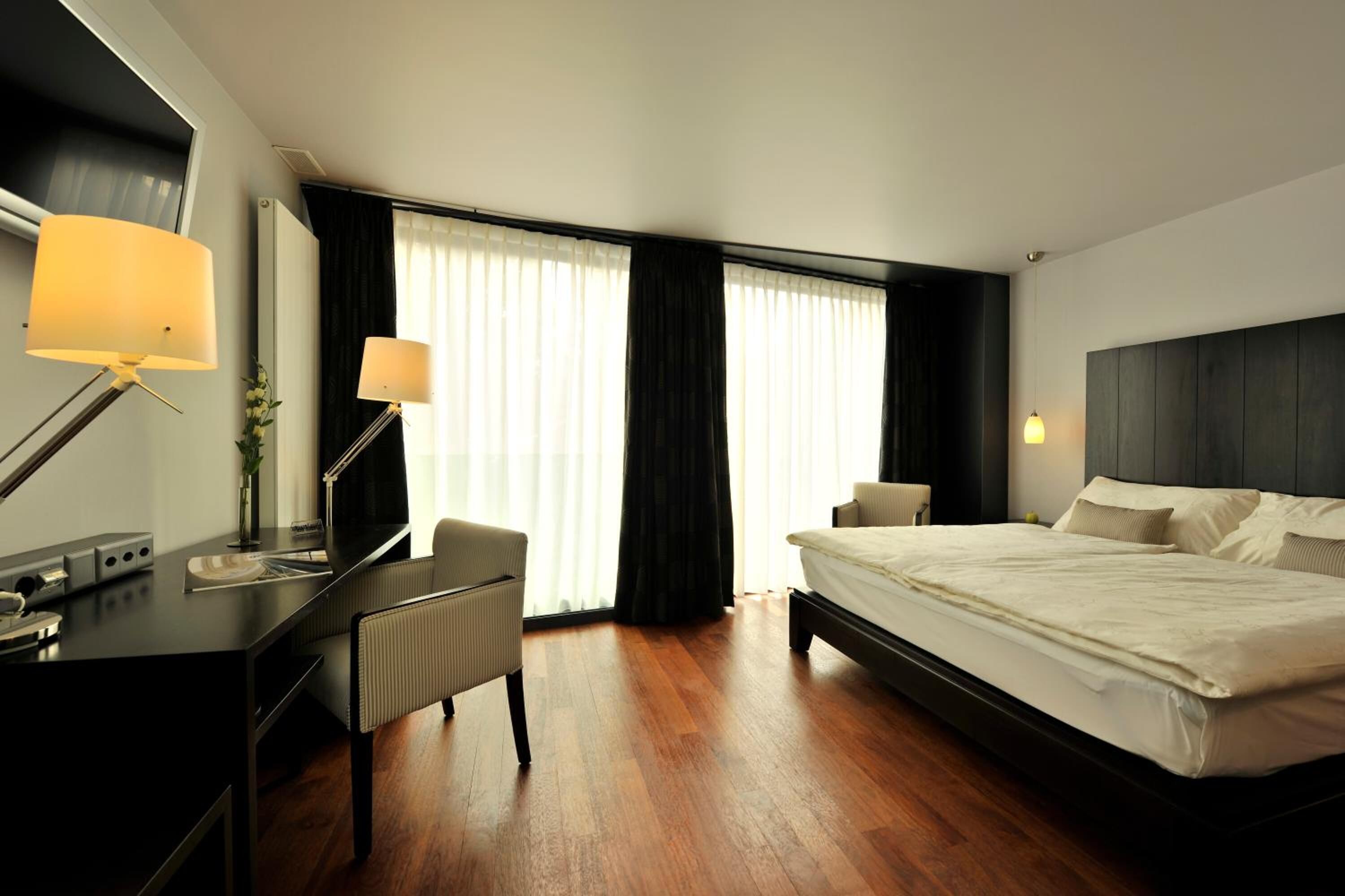 A hotel room with a bed, a desk, a chair, a lamp, and a window with curtains.