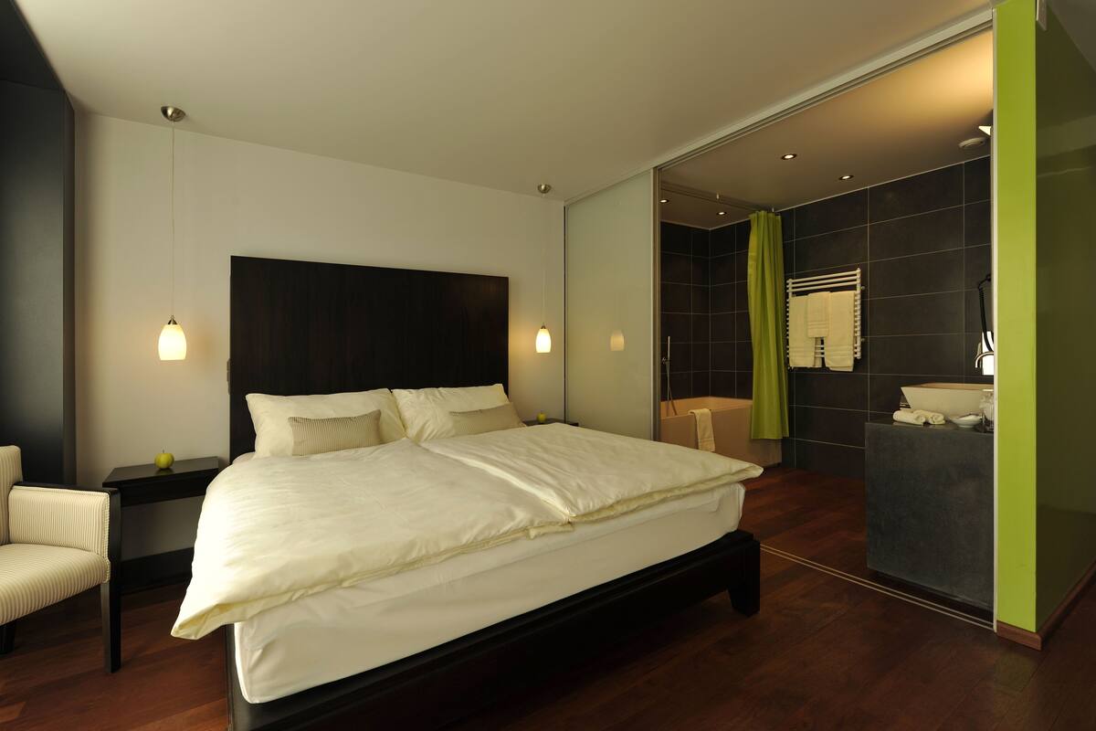 A modern bedroom with a large bed, bedside tables, a chair, and a bathroom with a sink and mirror.