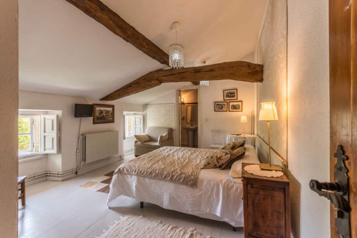 Ein Schlafzimmer mit einem großen Bett, Holzbalken, einer Chandelüster, einem Nachttisch mit Lampe und Blick ins Badezimmer.
