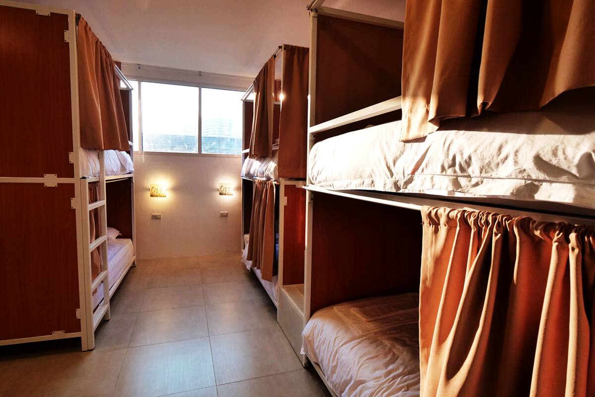A dormitory room with bunk beds, a window, and wall-mounted lights.
