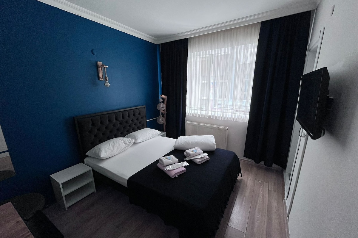 A hotel room with a bed, a TV, a window with curtains, a small shelf, and a radiator.