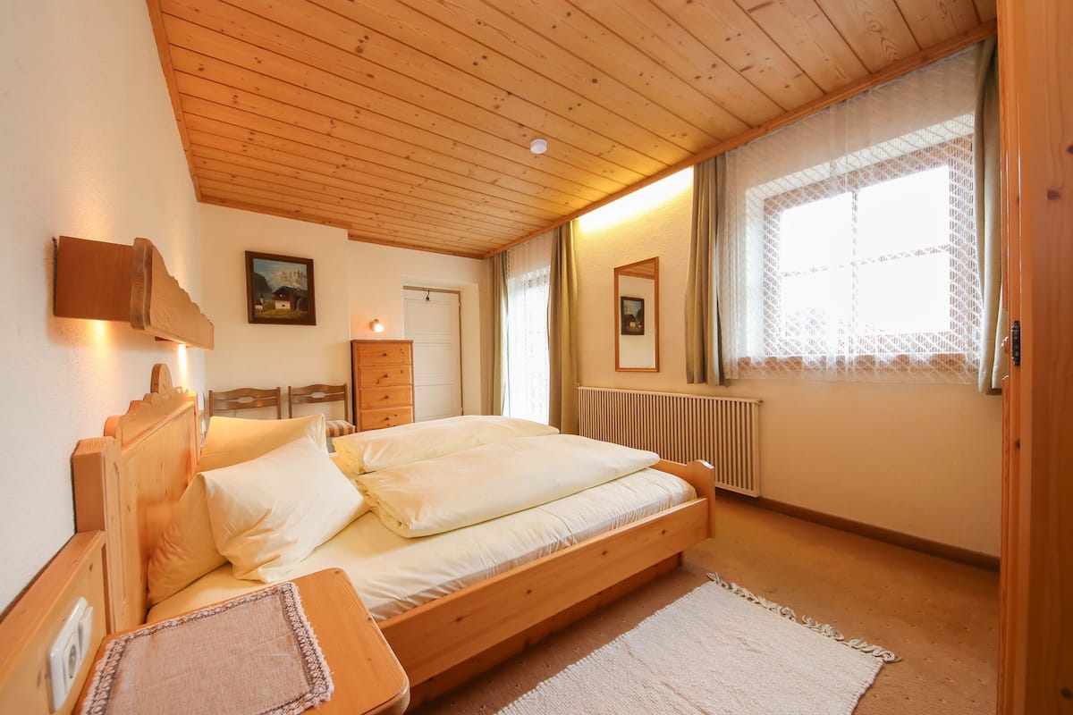 A bedroom with a wooden ceiling, a bed with white bedding, a nightstand, a dresser, and a window with curtains.