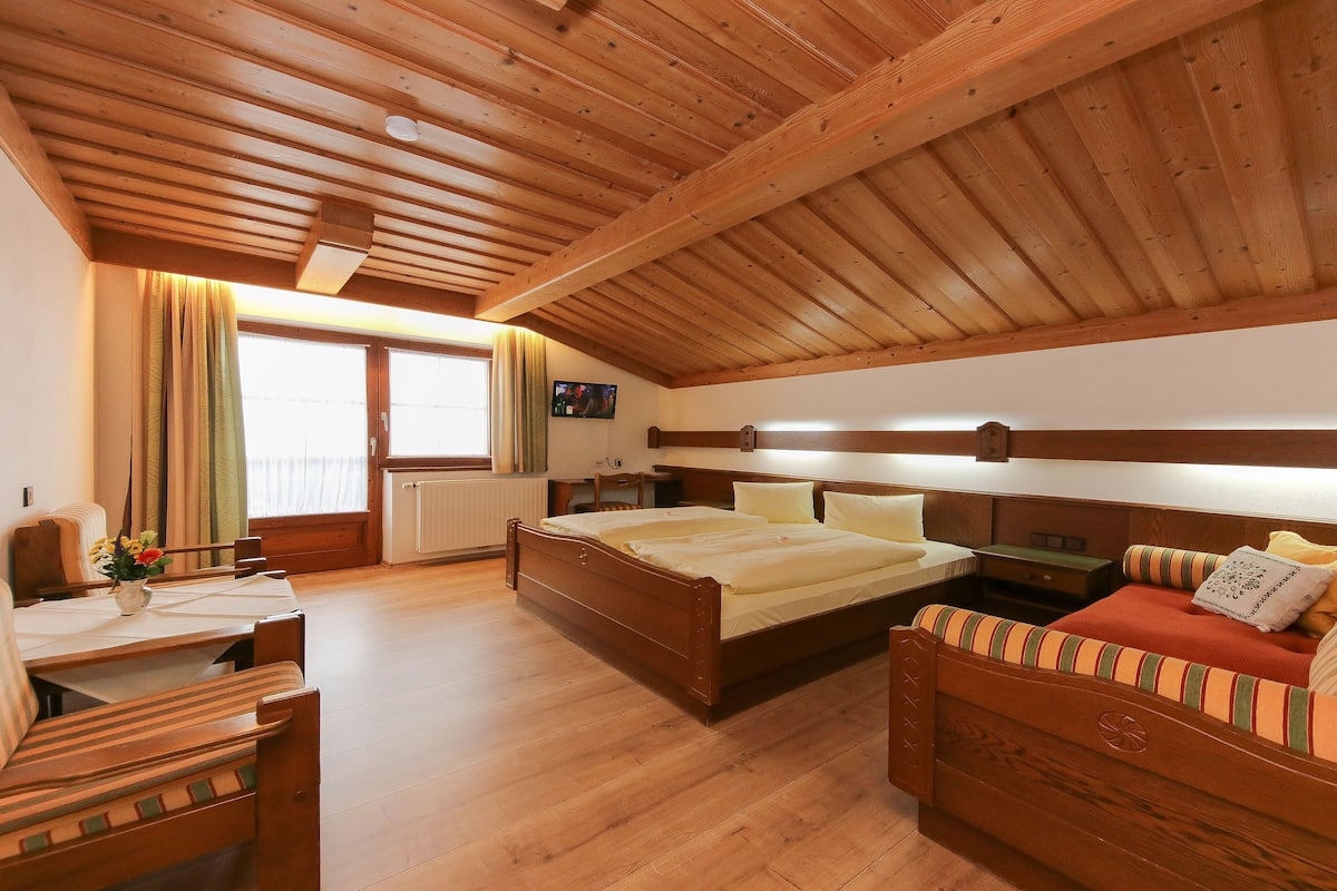 A wooden-floored bedroom with a bed, a sofa, a desk, and a window with curtains.