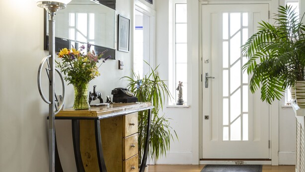 A hallway with a wooden floor, a desk with drawers, a standing lamp, and a large potted plant. - Twentieth Century B&B (Birchington)
