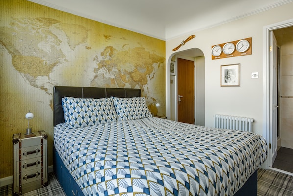 Un dormitorio con un mapa del mundo en la pared, una cama con ropa de cama estampada, un aparador con una lámpara y una puerta que da a otra habitación.