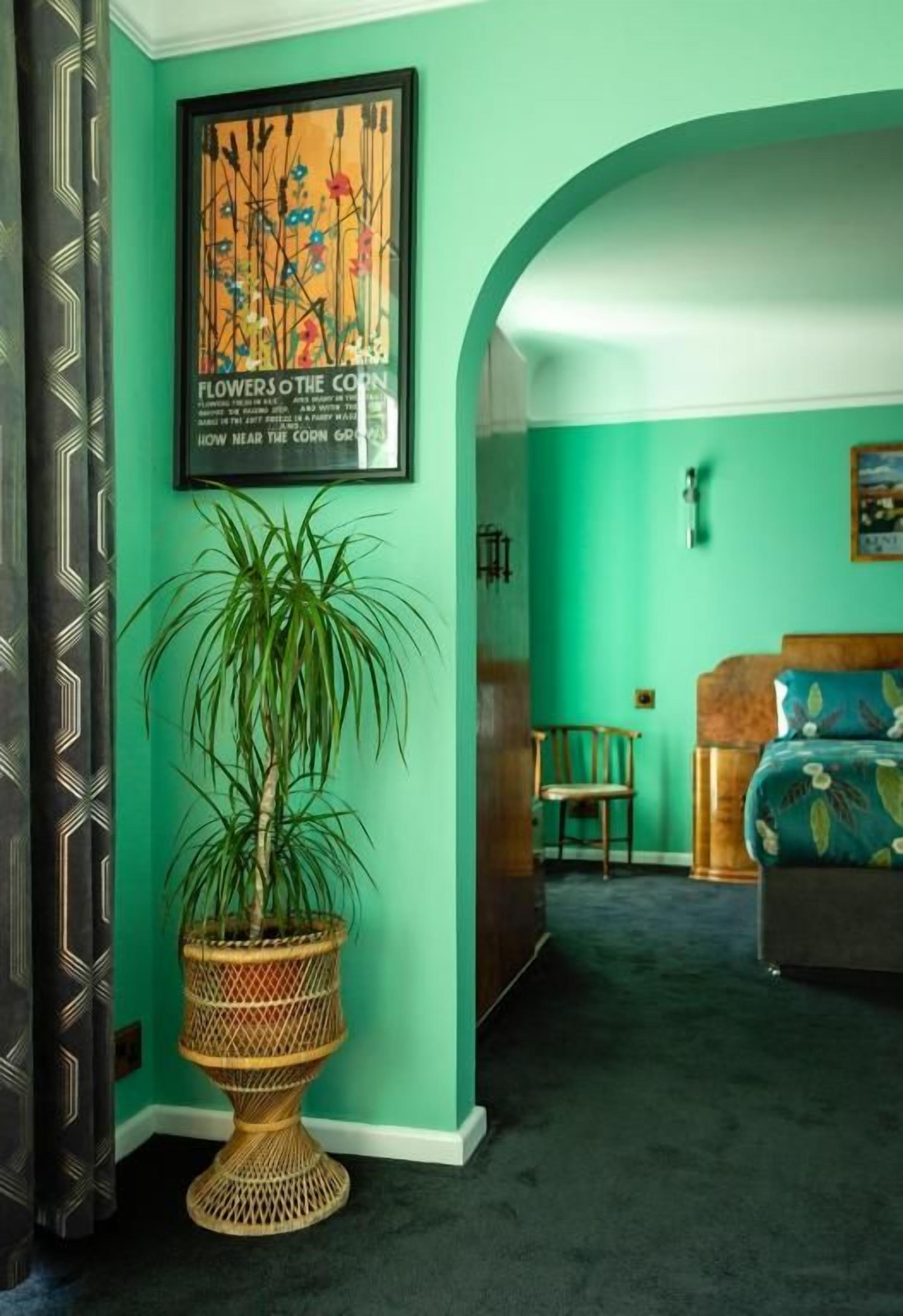 A room with a green wall, a large potted plant, a framed picture, and a bed with floral bedding.