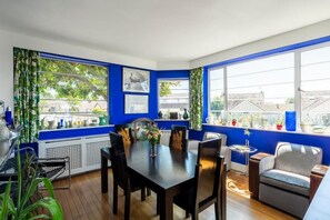 A dining area with a table and chairs, large windows with curtains, and a view of buildings outside. - Twentieth Century B&B (Birchington)