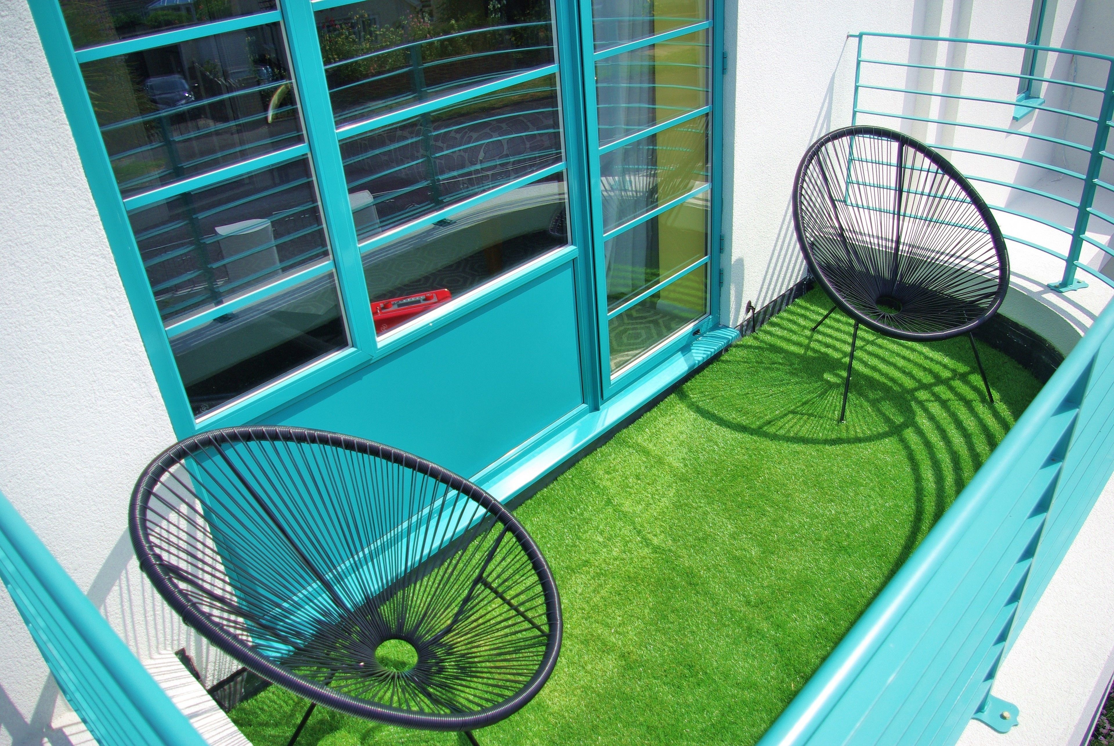 A balcony with artificial grass and two black wire chairs, a window with a blue frame, and a white wall.
