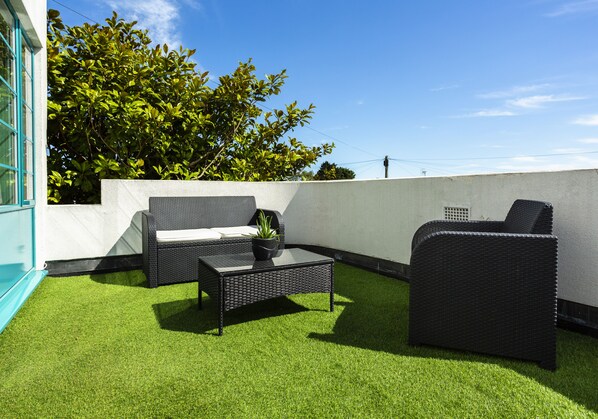 An outdoor patio with a sofa, chair, and coffee table, surrounded by artificial grass and a white wall. - Twentieth Century B&B (Birchington)