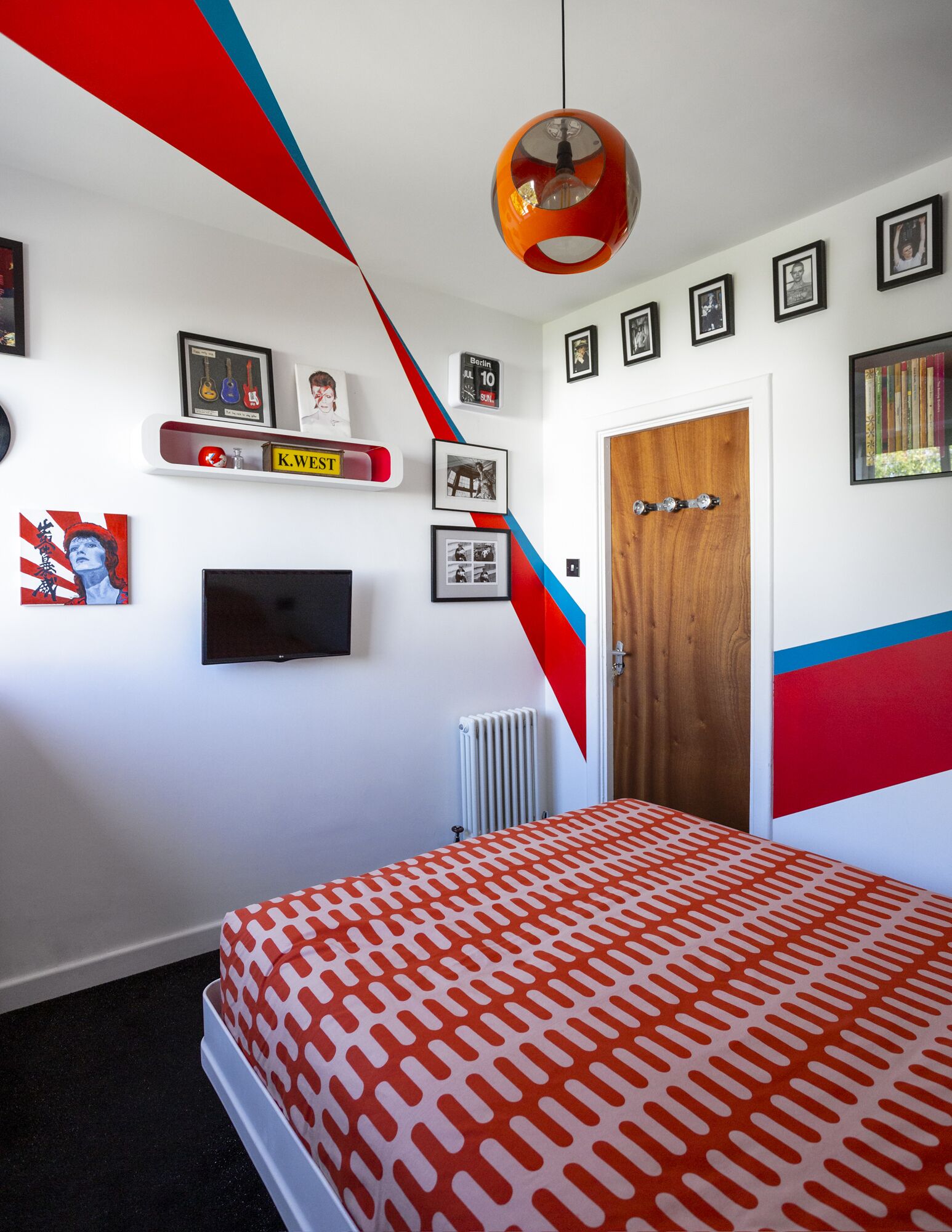 Un dormitorio con cama, televisión y radiador. Hay cuadros enmarcados en la pared y una puerta con manija.