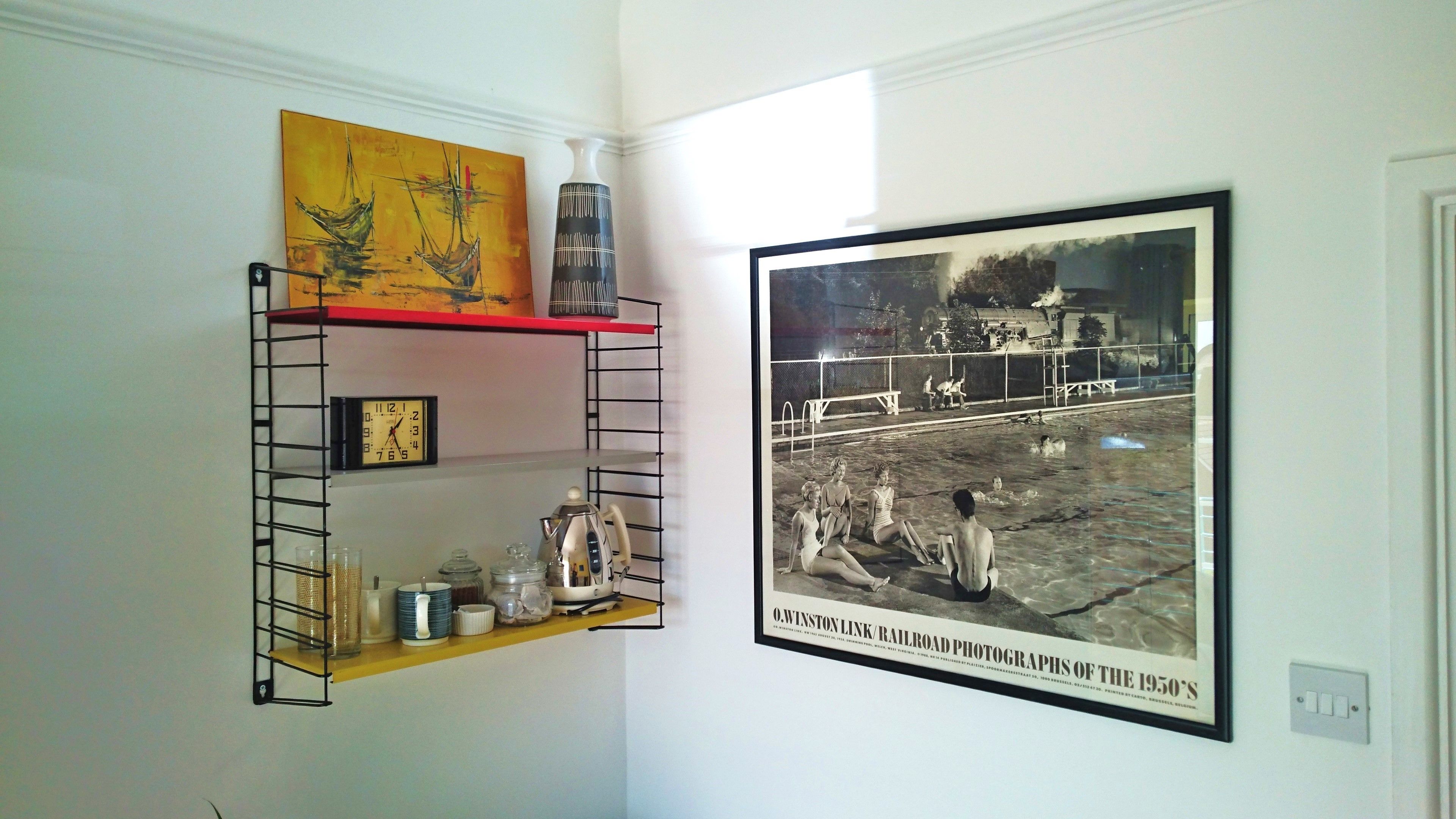A wall with a shelf holding a clock, jars, and a framed photograph of a swimming pool.