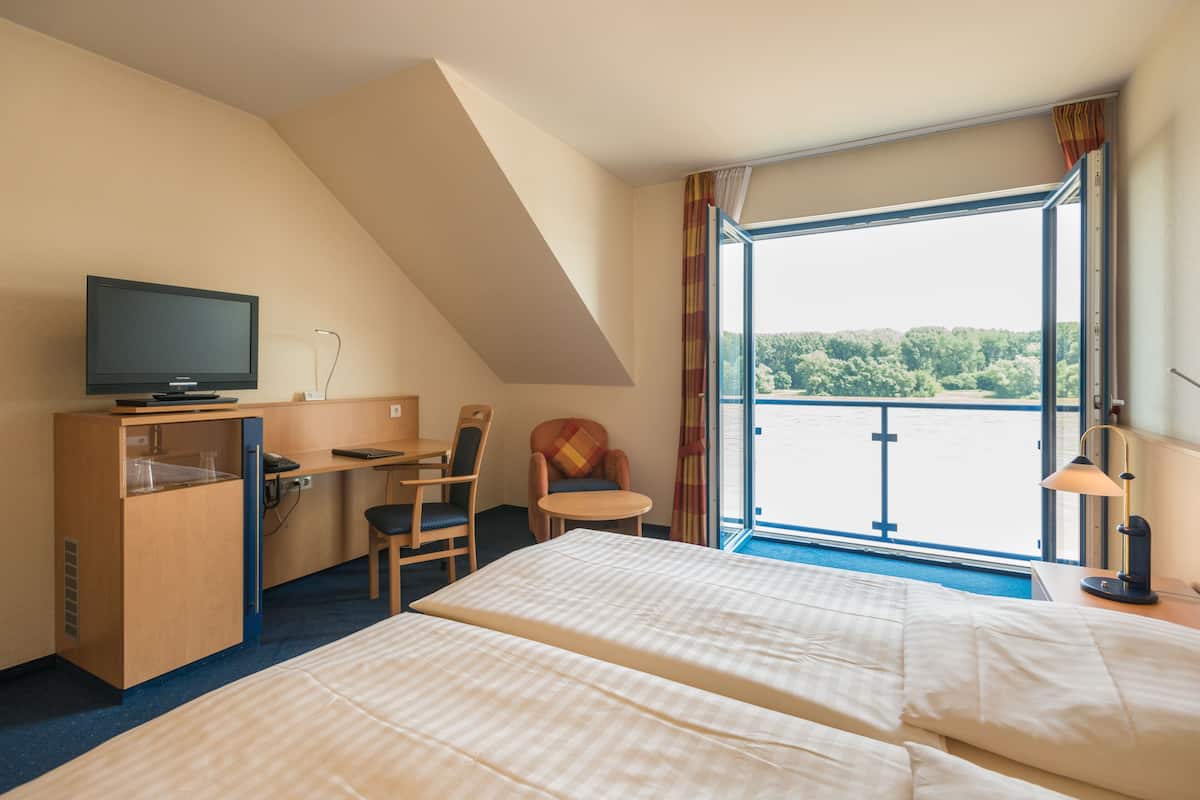 A hotel room with two beds, a desk with a television, a chair, a small table, and a window with a view of greenery.