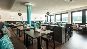 A modern restaurant with a high ceiling, large windows, and a central island. Tables and chairs are arranged around the island, and there are decorative plants and a menu board visible. - Rheinterrassen Hotel Café Restaurant (Bornheim)