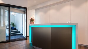 A reception desk with a blue backlight, a potted plant, and a staircase in the background. - Rheinterrassen Hotel Café Restaurant (Bornheim)