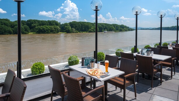 An outdoor dining area with wicker chairs and tables, a view of a river, and a boat in the distance. - Rheinterrassen Hotel Café Restaurant (Bornheim)