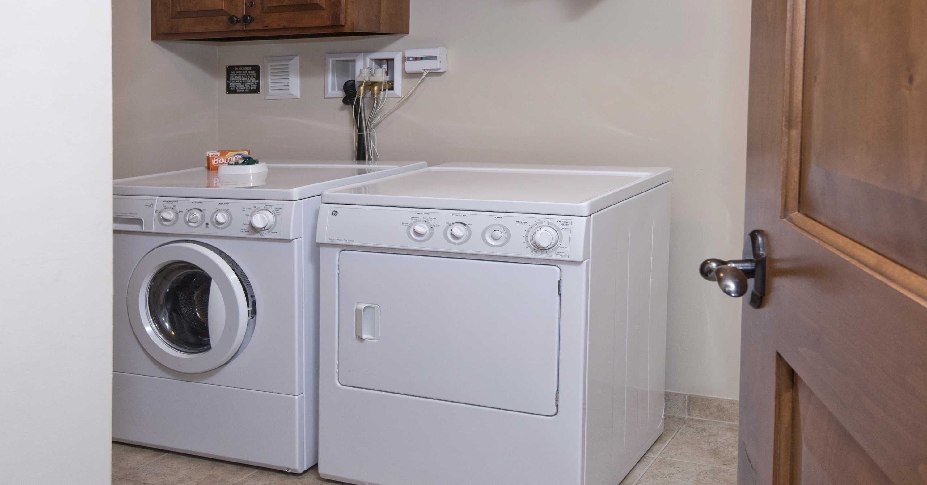 A laundry area with two white washing machines and a dryer, a wooden door, and a wall-mounted electrical outlet.