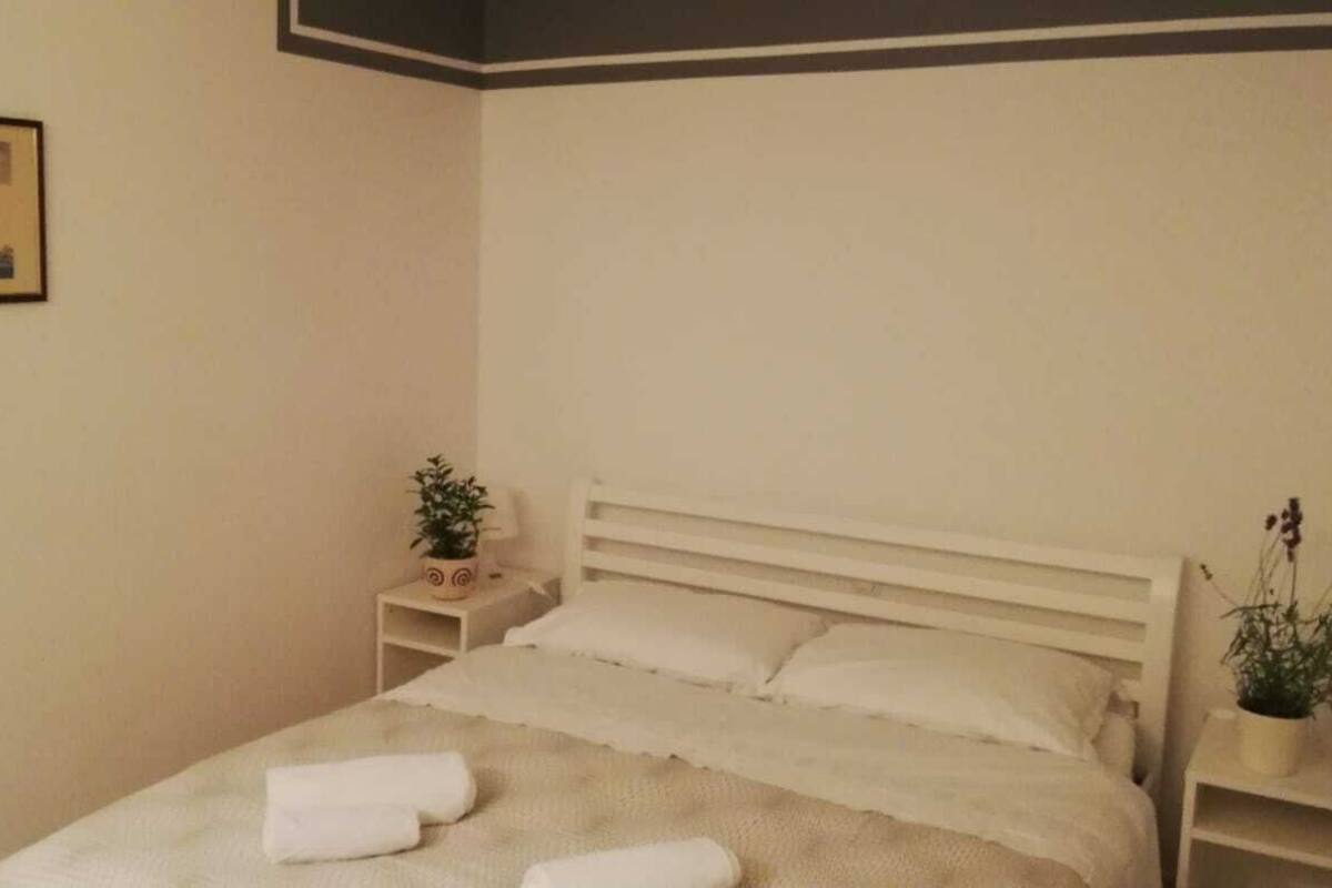 A neatly made bed with white linens and towels, a small bedside table with a plant, and a framed picture on the wall.