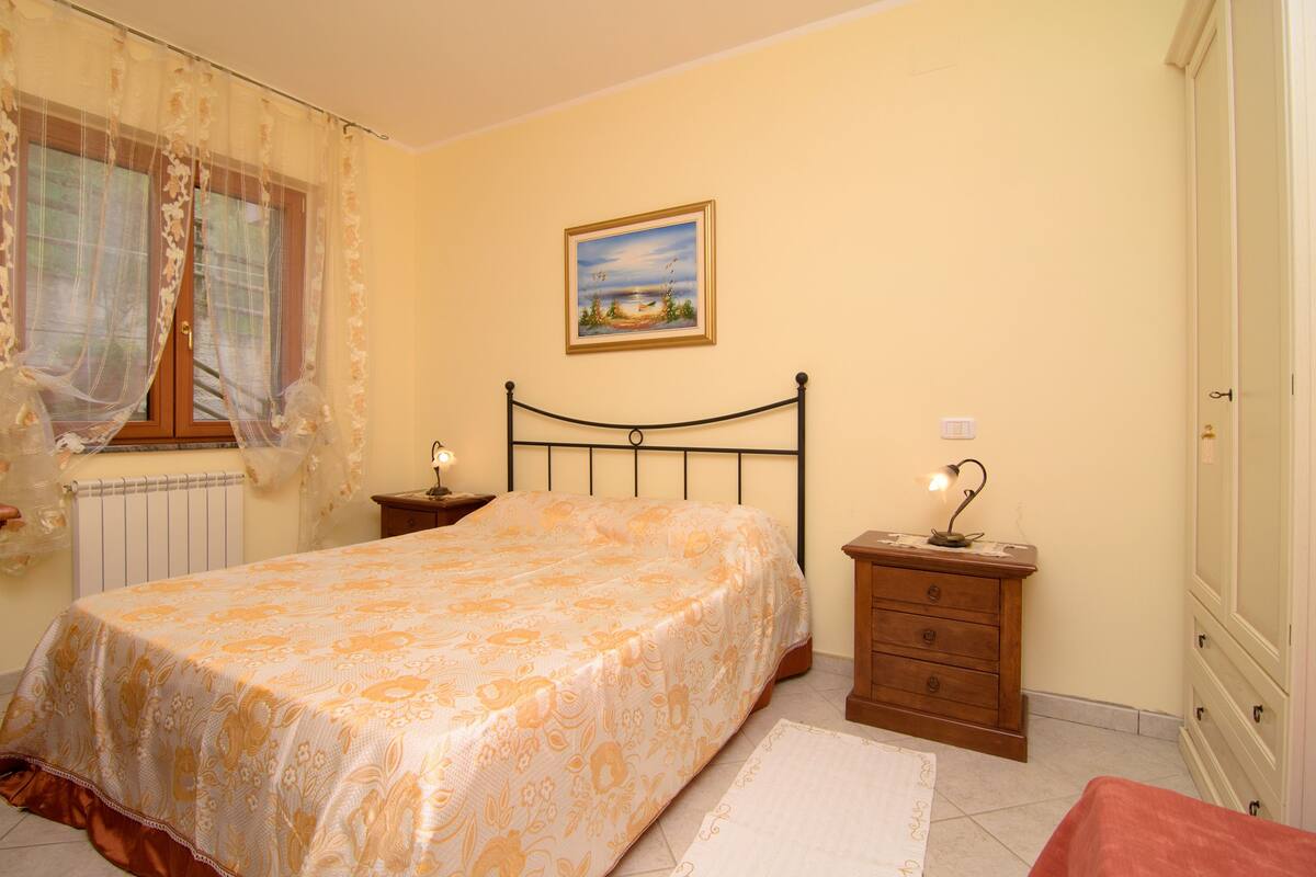 Une chambre à coucher comprenant un lit, une table de chevet, une commode, une fenêtre avec des rideaux et un tableau accroché au mur.