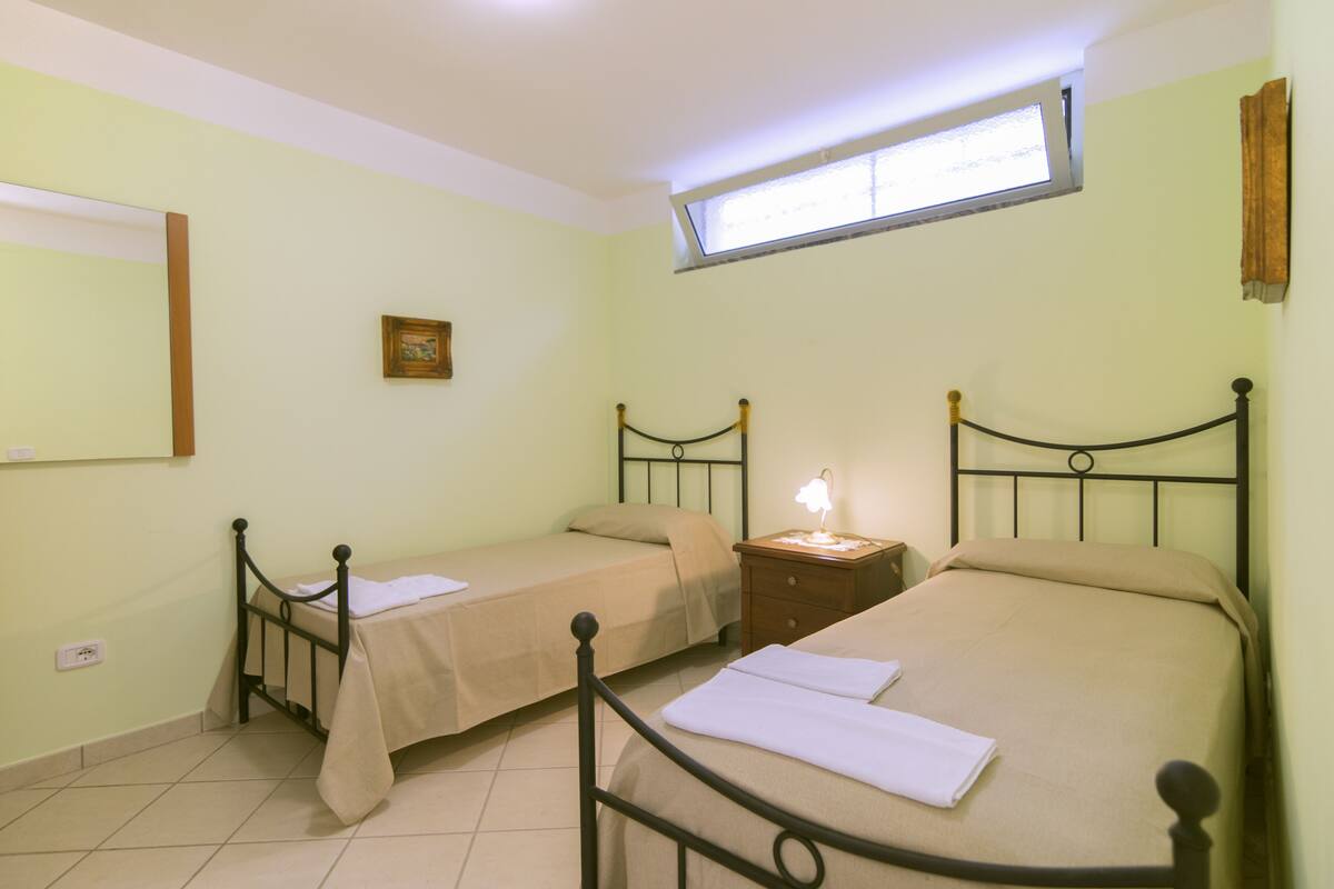 Une chambre avec deux lits simples, une table de chevet avec une lampe, une fenêtre et un tableau accroché au mur.