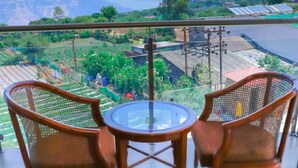 A balcony with two wicker chairs and a glass table, overlooking a green landscape with solar panels and buildings. - Bliss County Resort (Mahabaleshwar)