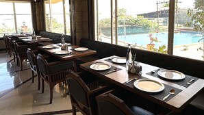 A restaurant with wooden tables and chairs, large windows with a view of a pool and greenery. - Bliss County Resort (Mahabaleshwar)