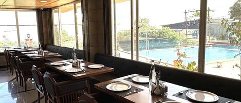 Un restaurante con mesas y sillas de madera, ventanas amplias que dan a una piscina y follaje.