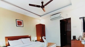 A hotel room with a large bed, a wooden headboard, a ceiling fan, an air conditioning unit, a wooden cabinet, a desk with a chair, and a framed picture on the wall. - Bliss County Resort (Mahabaleshwar)