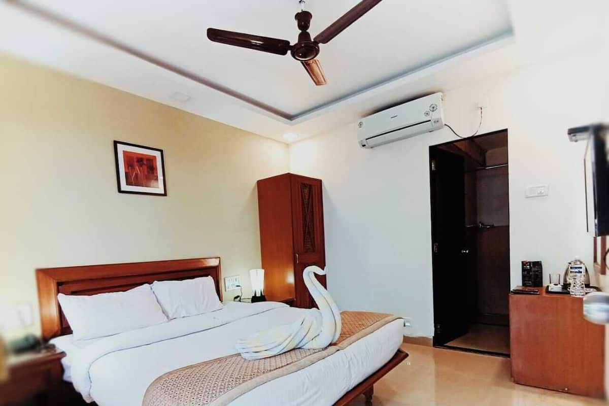 A hotel room with a large bed, a wooden headboard, a ceiling fan, an air conditioning unit, a wooden cabinet, a desk with a chair, and a framed picture on the wall.