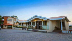 A hotel with a modern design, featuring a central building with a flat roof and a side wing. There are outdoor seating areas and a paved entrance. - Bliss County Resort (Mahabaleshwar)