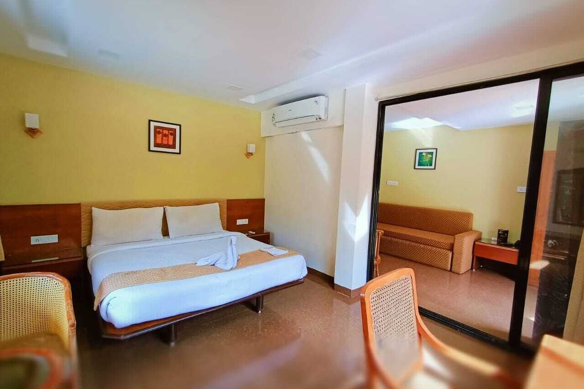 A hotel room with a bed, a sofa, a chair, an air conditioning unit, and a door leading to another room.