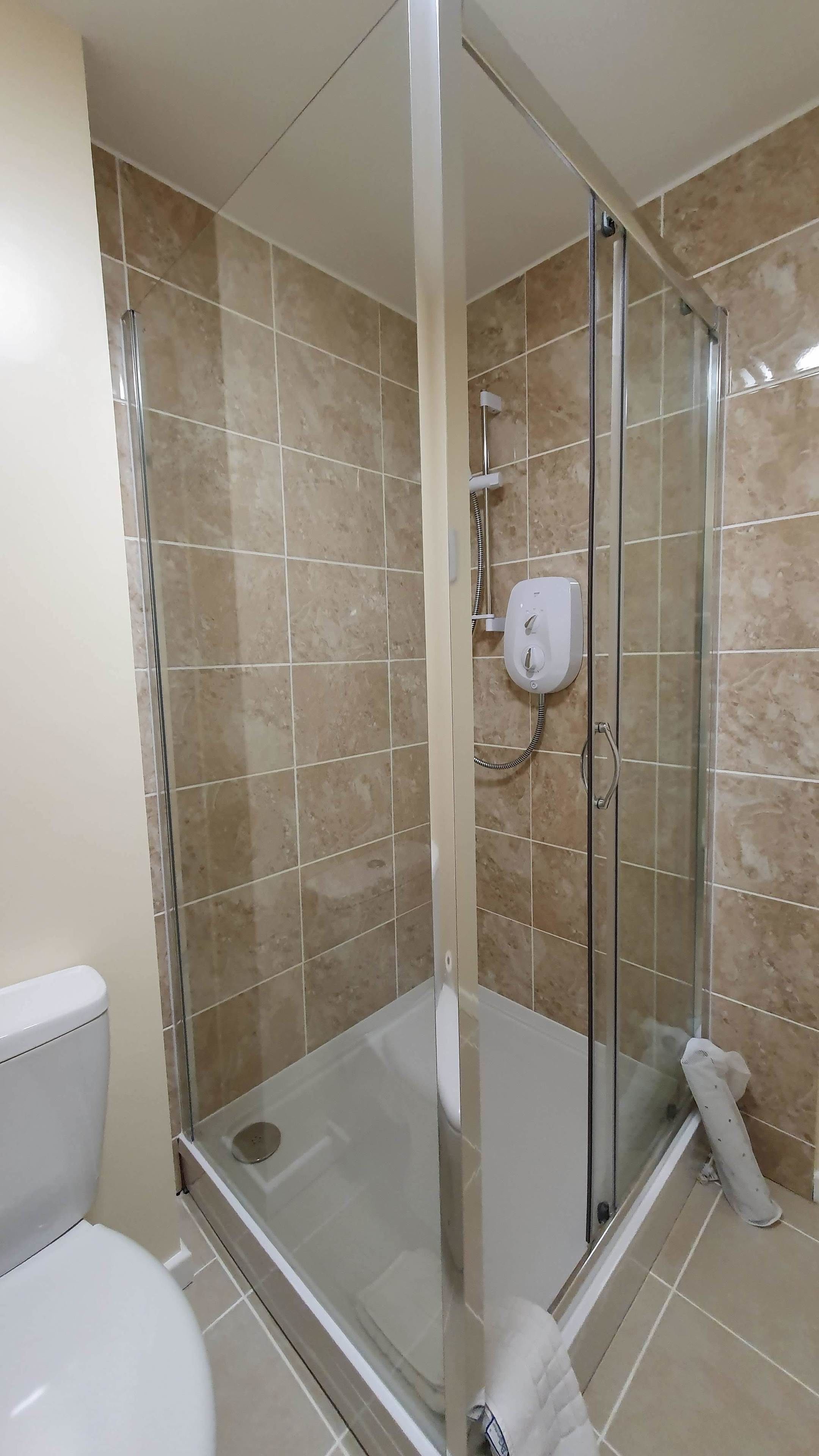 A glass-enclosed shower with a handheld showerhead, a white toilet, and beige tiled walls.