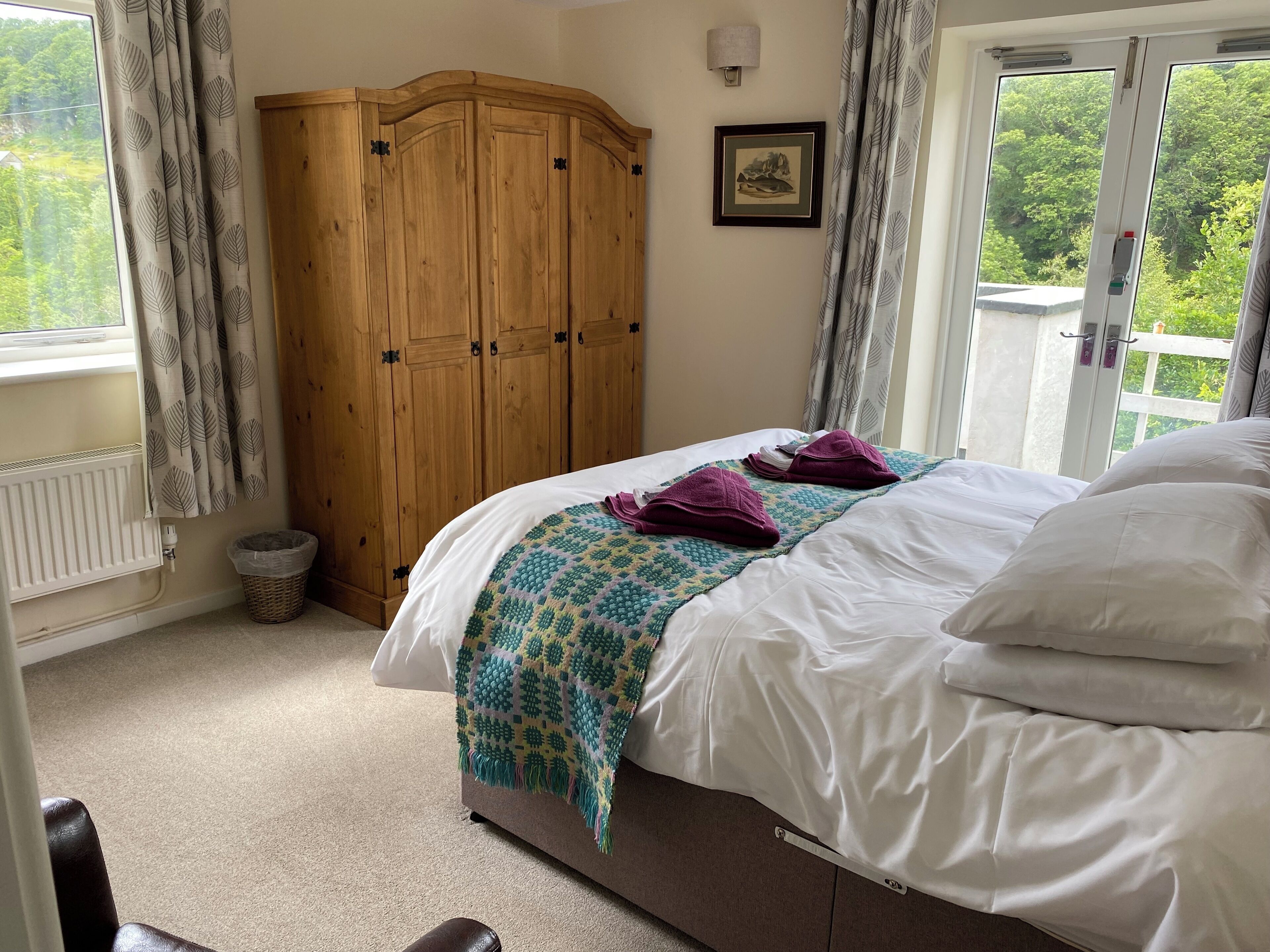A bedroom with a bed, a wooden wardrobe, a radiator, a window with curtains, and a view of greenery outside.