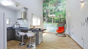 A modern kitchen with a central island, bar stools, a hanging egg chair, and a large window with a forest mural.