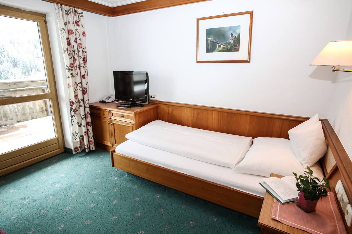 Una habitación con una cama de madera, una mesita de noche con lámpara, televisi ón y una ventana con cortinas florales.