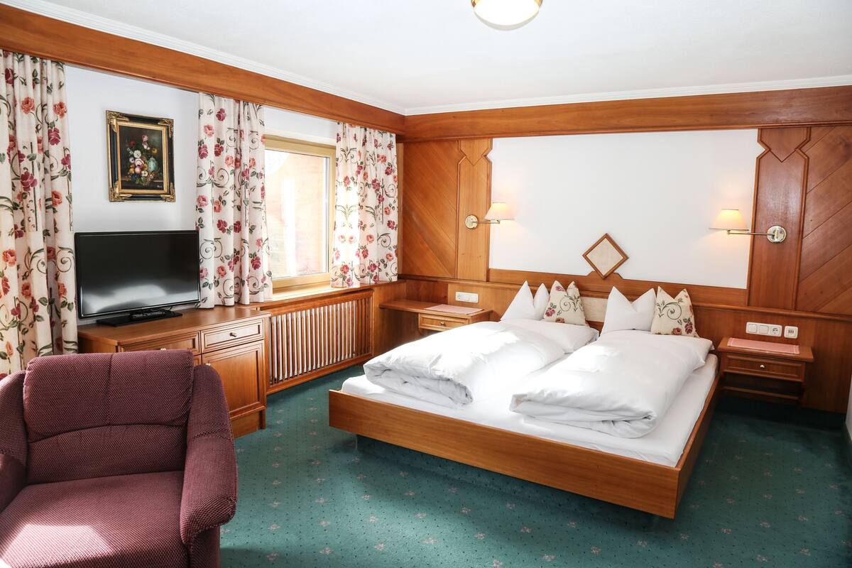 Una habitación de hotel con una cama de madera, un televisor, un sillón rojo y cortinas con estampado floral.