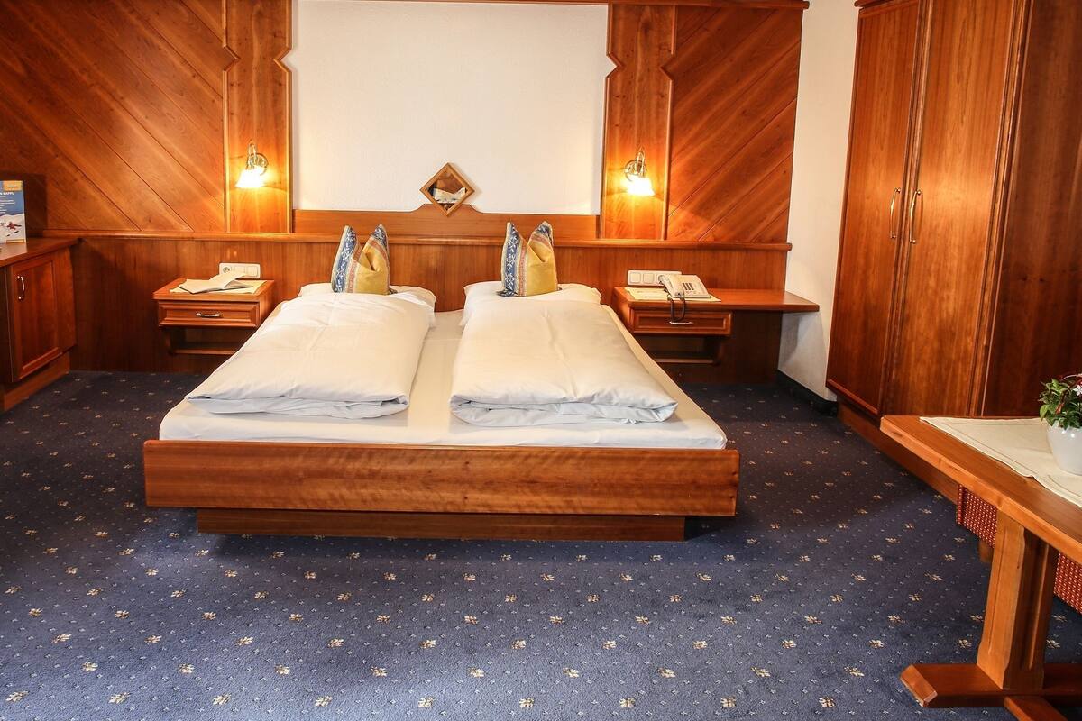 Una habitación de hotel con una cama grande, cabecera de madera, dos mesitas de noche con lámparas, un escritorio con silla y una planta pequeña en una mesita auxiliar.