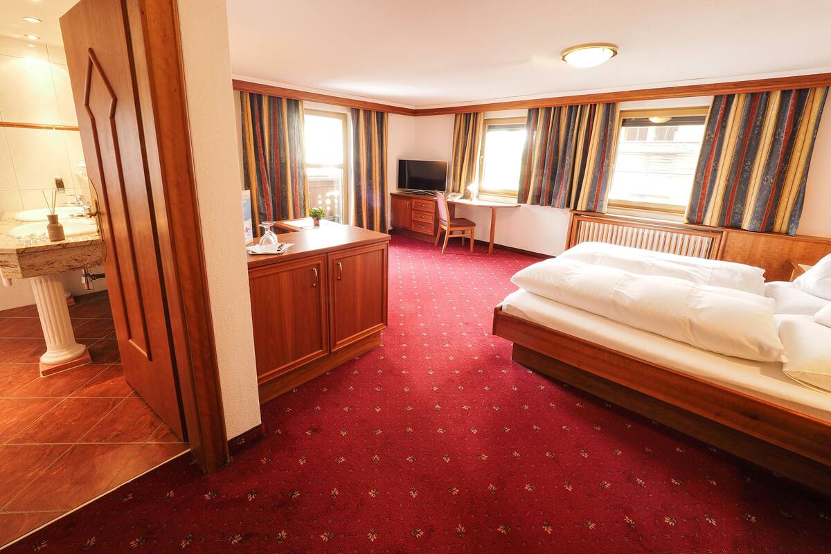 Habitación de hotel con cama, escritorio, silla, televisor y baño con lavamanos y espejo.