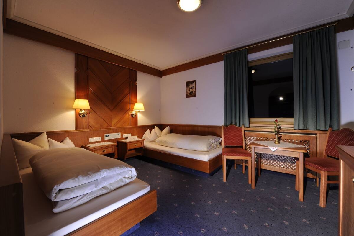 Habitación de hotel con cama, mesitas de noche, escritorio con silla, ventana con cortinas y una lámpara de pared.
