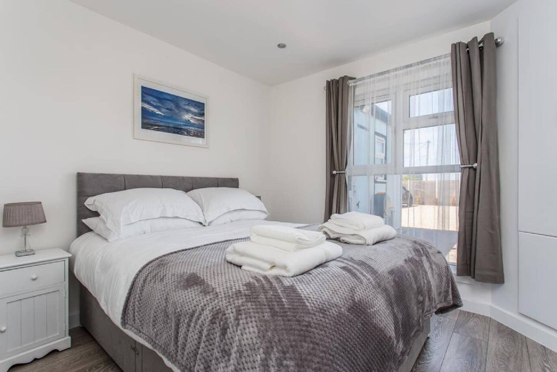 A bedroom with a bed, a nightstand, a lamp, a window with curtains, and a painting on the wall.