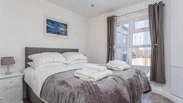 A bedroom with a bed, a nightstand, a lamp, a window with curtains, and a painting on the wall.