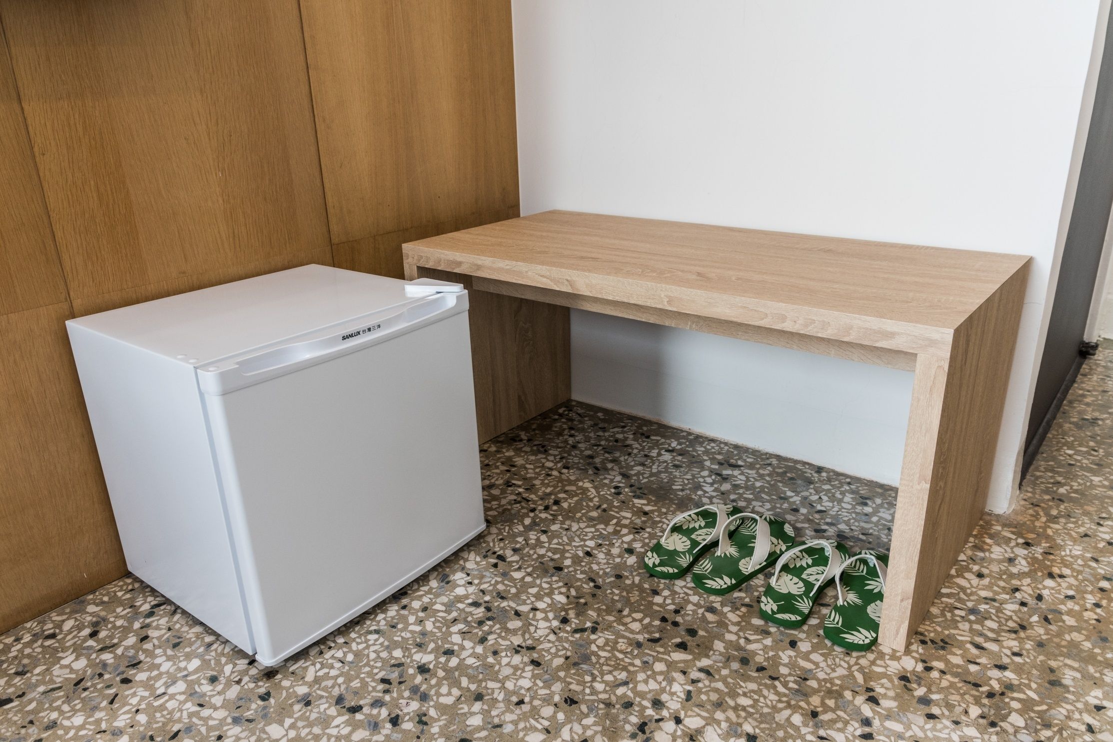 Un refrigerador blanco y compacto junto a un banco de madera sobre un suelo moteado.