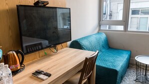 Ein Zimmer mit Holztisch, einem blaugrünen Sofa, einem an der Wand befestigten Fernseher und einem Fenster mit Blick auf Gebäude.